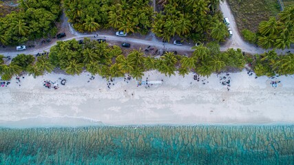 Aerial view of a tropical beach with palm trees, parked cars, and clear turquoise water