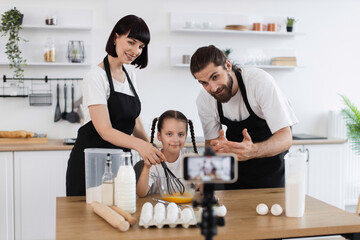 Caucasian family including adult man, woman, and child creating a cooking vlog in kitchen. Smiling...