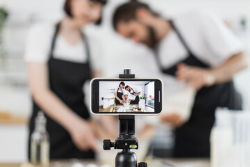 Caucasian parents and child, dressed in aprons, knead dough for video blog in home setting. Emphasizes modern content creation, family collaboration, and homemade culinary activities