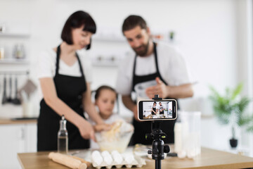 Caucasian family of two adults and one child recording cooking video preparing dough. Scene...