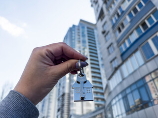 A woman's hand holds a house key in plain view in front of a tall, elegant modern building rising