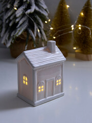 Ceramic house ornament emits a warm glow in a festive setting with string lights and snow-covered...