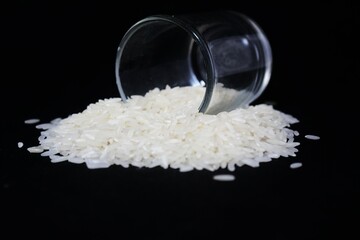A small, clear glass is tilted to the side against a pitch-black background. The cup is filled with a pile of raw, white rice, which is piled up in front of the cup. The rice grains are long and shiny