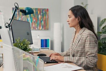 An attractive, experienced company accountant sits in office in front of a computer checking...