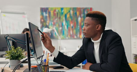 Handsome dark-skinned man, corporate office worker spends morning in front of computer, points at screen, counts, ponders, checks correctness of graphs, completes data