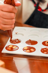 A person uses a piping bag to fill chocolate molds, with some containing nuts. This process showcases homemade chocolate making, perfect for culinary enthusiasts
