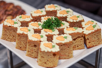 The image features square carrot cakes topped with orange and white frosting styled as carrots, garnished with parsley at the center. The cakes are neatly arranged, presenting an inviting dessert