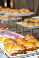 The image displays a variety of food, focusing on sandwiches made with sesame seed buns, lettuce, tomato, and other fillings. In the background, there are trays of colorful pastries, adding vibrancy