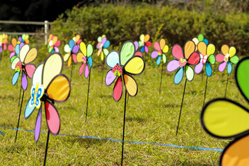 A lively field of colorful pinwheels spins on green grass, enhanced by butterfly and ladybug decorations. Bright hues and a playful vibe are framed by bushes and a charming fence