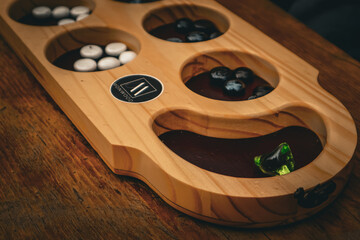The image depicts a wooden Mancala board by MOONWOOD, with circular compartments holding black, white, and one green glass piece. It appears as a strategy tabletop game for two players