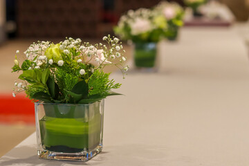 A small floral arrangement featuring green leaves, delicate white baby's breath, and a light green rose, presented in a square glass vase with layered green material at the base