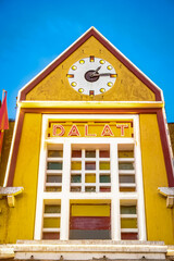 A building facade featuring a large clock with a white face and black hands above the bold red letters spelling 'DALAT,' on a yellow background with white and red accents