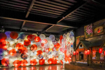 A decorative wall adorned with a variety of traditional Japanese umbrellas in vibrant colors and intricate patterns, predominantly red and white. A small structure with glowing red lanterns complement