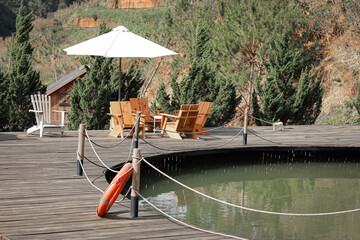 A serene wooden deck featuring a table and chairs under a white umbrella, overlooking a tranquil pond. A lifebuoy is attached to a post nearby, surrounded by lush greenery and a small wooden structure
