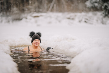 Fototapeta premium Smiling woman in black swimwear and winter hat enjoys an ice bath in a snowy outdoor setting, surrounded by frozen landscape and falling snowflakes..