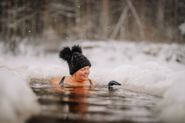 Smiling woman in a black swimsuit, hat, and gloves enjoys cold water immersion in a snowy ice hole...