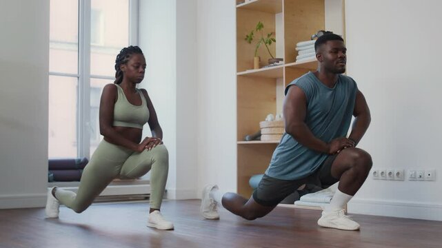 Muscular Black fitness instructor explaining how to perform lunge stretch while leading personal workout with young woman in modern minimalist gym