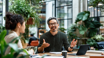 An Asian businessman engages in a discussion with a colleague in a modern office filled with plants.