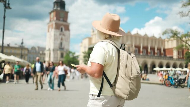 Elderly woman exploring the city center with a phone in her hand, enjoying the sights. She is wearing a hat and a backpack, walking in a popular tourist spot on a sunny day