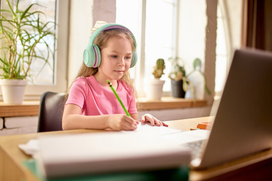Curious girl in pink shirt wearing mint headphones writing in notebook while attentively watching laptop screen during online class at home. Concept of childhood, online education, remote studying