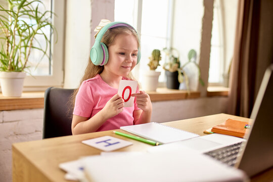 Curious girl leaning on desk and watching laptop screen with teacher leading lesson attentively during online class. Child studying at home in well lit room. Concept of education, childhood