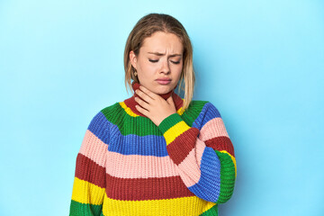 Blonde young woman in multicolor sweatshirt on blue background suffers pain in throat due a virus...
