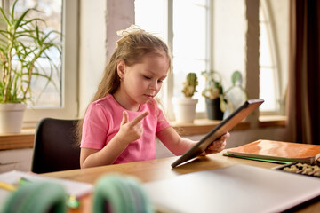Girl sitting at desk, counting with fingers, using tablet for online lesson. Child studying...