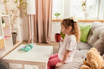 Little girl taking cozy break with red mug after online lesson, sitting on couch with headphones on...