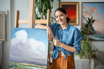 Portrait of accomplished female painter smiles and presenting freshly completed landscape artwork painting. Workshop with landscape artworks, masterclass session, smiling teacher ready to work