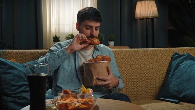 Man spending time at home with fast food delivery, sits on couch watching tv, dipping fried chicken leg in sauce and biting it, enjoying the taste.