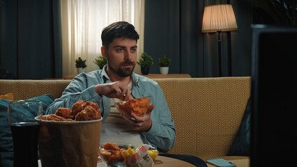 Bearded man spending time at home with fast food delivery, sits on couch watching tv and eating...