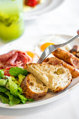 Close-up of a breakfast plate with salad, bread, sausages, bacon, sunny-side-up eggs, and a green drink in the background. Fresh, colorful, and appetizing.