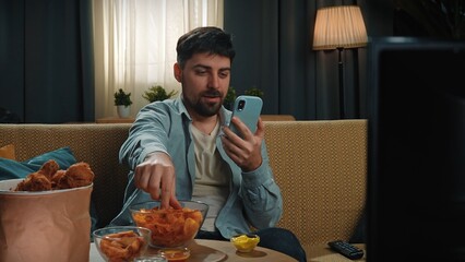 Bearded man spending time at home with fast food delivery, sits on couch, watching tv eating potato...