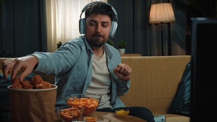 Bearded man spending time at home with fast food delivery, sits on couch listening music in headphones on smartphone and eating fried chicken.