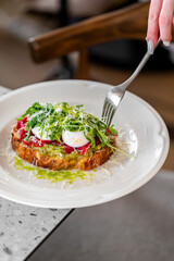 A close-up of a gourmet dish featuring toast topped with fresh greens, cherry tomatoes, poached eggs, and grated cheese, served elegantly on a white plate