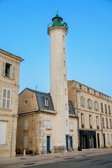 Fototapeta premium An old lighthouse proudly stands near historic harbor buildings, surrounded by calm waters and under a clear blue sky, creating a picturesque scene that embodies maritime history and charm.