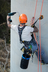 Construction worker using drill while hanging on ropes on concrete wall