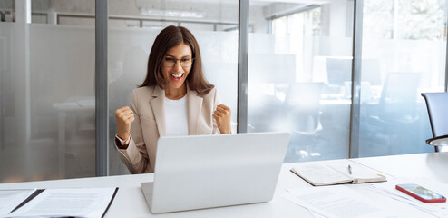 Emotional business woman showing happiness getting bonus reward or good news on screen laptop pc computer in office. Excited young businesswoman winner showing triumph yes gesture. Banner. copy space
