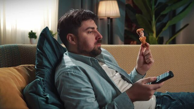 Bearded man spending time at home with fast food delivery, lays on couch, watching tv switching channels and eating fried chicken.
