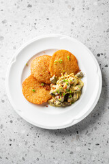 Crispy golden potato pancakes served on a white plate alongside a creamy mushroom salad, garnished with green herbs on a textured gray background.