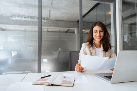 Smiling latin business woman saleswoman working on laptop computer reading financial contract document in office workplace. Young manager businesswoman specialist doing paperwork using pc. Copy space