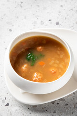 A bowl of clear soup with meatballs, diced carrots, onions, and green vegetables on a white plate, placed on a speckled gray countertop, perfect for culinary presentations.