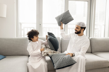 Cheerful Arabic son and daddy spend carefree pastime together in cozy living room, playing seated...