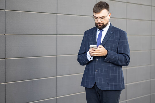 businessman with beard and glasses stands attentively against grey wall. He is dressed in a tailored blue suit with a checked pattern and a tie, focusing on his smartphone. concept: using a phone