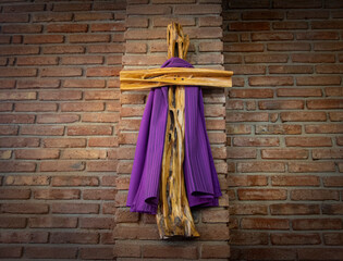 A cross made of distressed wood draped in purple for observance of Lent. A brick wall is in the...