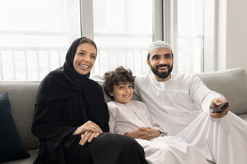 Arabic couple with little son dressed in traditional attire relaxing on couch, holding remote...