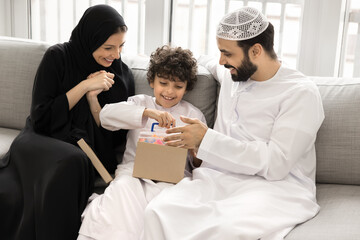 Adorable little 7s boy opening parcel box with gift while sitting on couch with loving parents, family dressed in traditional attire congratulate son with birthday or life event. Bonding, celebration