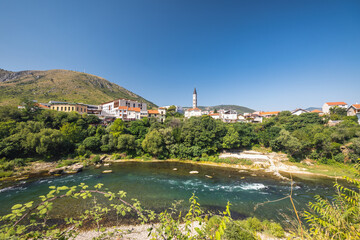 Fototapeta premium Neretva river in Mostar town, Bosnia and Herzegovina, Europe. Scenic view of a town nestled along a riverbank, featuring charming architecture and lush greenery under a clear blue sky. Peaceful.
