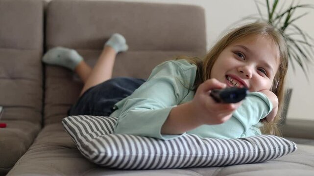 Caucasian child girl laying on sofa home and watching tv,using pilot,remote control.Happy kid indoors have a fun.Lesure concept. 
