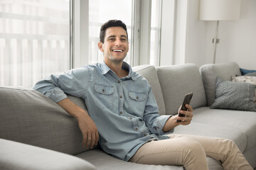 Cheerful young man sitting on sofa in cozy living room, holding his smartphone, smiling at camera,...